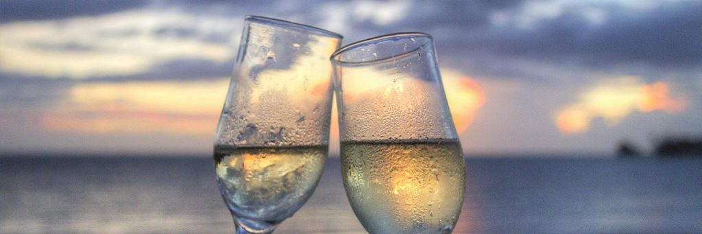 Two wine glasses with light yellow liquid clinking together with a sunset beach behind