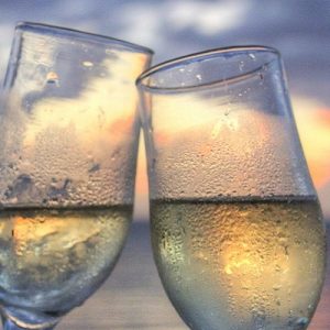 Two wine glasses with light yellow liquid clinking together with a sunset beach behind