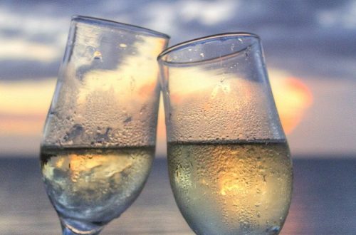 Two wine glasses with light yellow liquid clinking together with a sunset beach behind