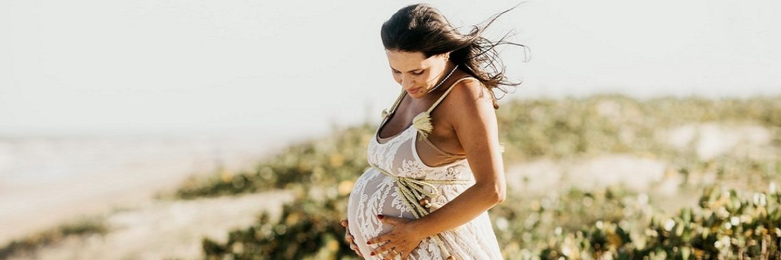 Pregnant woman in a flowing white dress holding her belly while relaxing on the beach