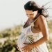 Pregnant woman in a flowing white dress holding her belly while relaxing on the beach