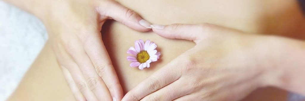 Woman's hands resting on her belly in the shape of a love heart with a small pink flower on her bellybutton