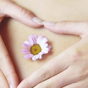 Woman's hands resting on her belly in the shape of a love heart with a small pink flower on her bellybutton